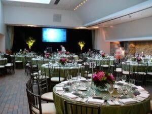 An elegant banquet hall setup with round tables covered in green linens, surrounded by dark wooden chairs. Each table is adorned with vibrant floral centerpieces and complete table settings, including wine glasses and folded napkins. A large screen and subtle lighting enhance the ambiance of the spacious venue.