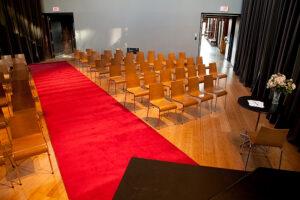 A minimalist indoor ceremony setup featuring rows of wooden chairs arranged on either side of a red carpet aisle. The space has a modern design with polished wood floors, neutral walls, and simple floral accents on a side table, creating an elegant and intimate atmosphere.