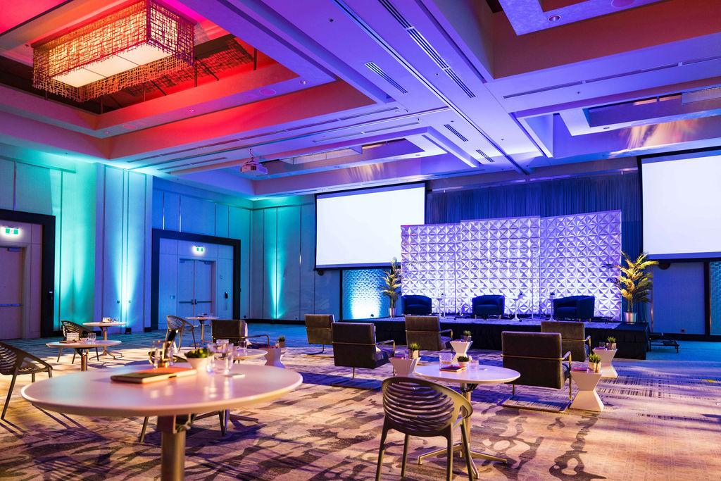 Design Inspiration 82 Modern conference and event space with colorful ambient lighting, presentation screen, cocktail tables, and lounge seating arranged for a corporate gathering.